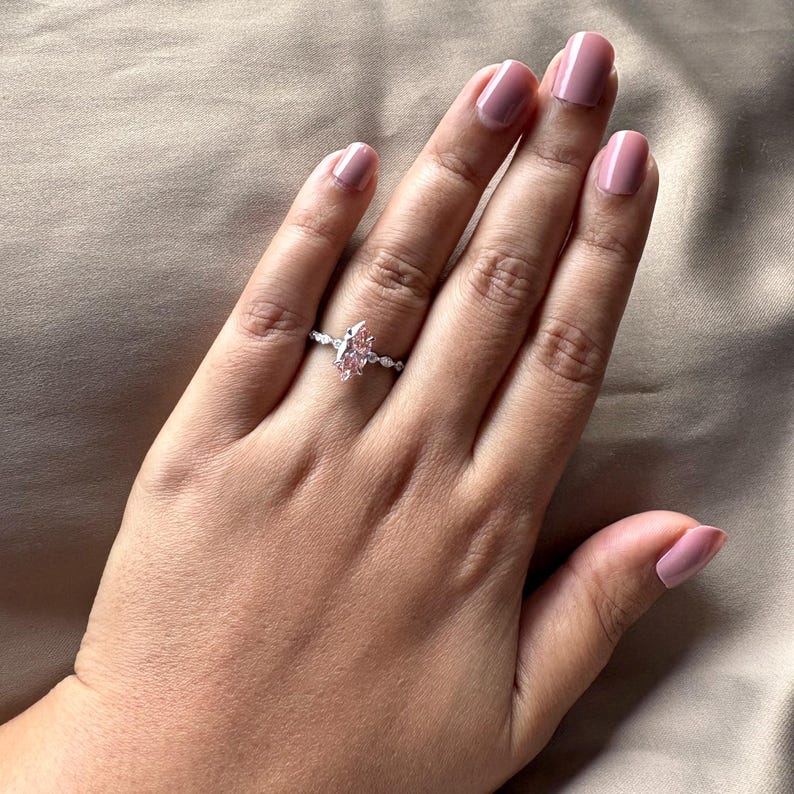 May include: A silver ring with a pear-shaped, light pink gemstone as the centerpiece. The band is adorned with small, clear stones. The ring is displayed on a hand with matching pink nail polish.