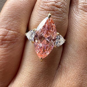 Pink marquise lab grown diamond engagement ring with heart-shaped side stones, three-stone design, 4.00 carat center diamond, elegant wedding jewelry.