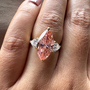 Pink marquise lab grown diamond engagement ring with heart-shaped side stones, three-stone design, 4.00 carat center diamond, elegant wedding jewelry.