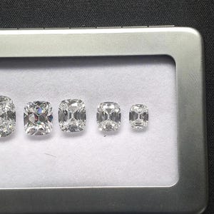 May include: Five clear, faceted cushion-cut gemstones arranged in a row from largest to smallest inside a rectangular silver-colored metal display tin with a clear window and a white fabric lining, showcasing brilliant light reflection.