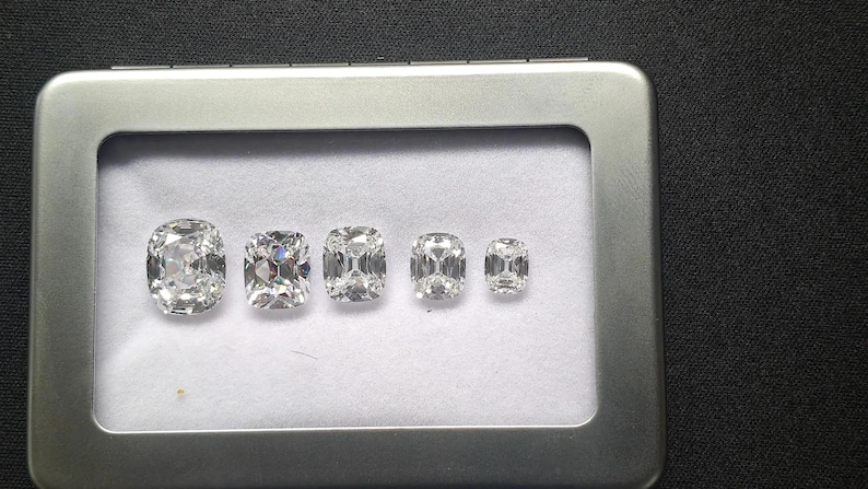 May include: Five clear, faceted cushion-cut gemstones arranged in a row from largest to smallest inside a rectangular silver-colored metal display tin with a clear window and a white fabric lining, showcasing brilliant light reflection.