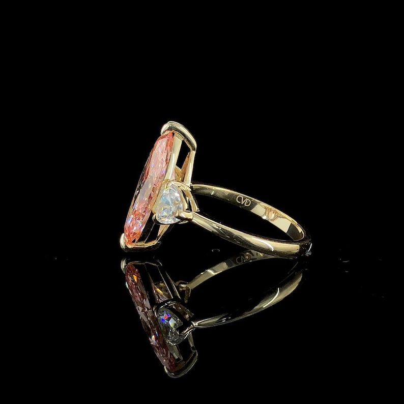 May include: A gold ring featuring a large, light orange, marquise-cut gemstone. A smaller, white, pear-shaped stone is set on one side. The ring is set against a black background, with the ring's reflection visible.