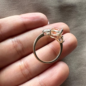 May include: A gold ring featuring a large, peach-colored gemstone flanked by two smaller, clear stones. The ring's band is a simple, polished gold, and the setting highlights the central stone's shape and color.