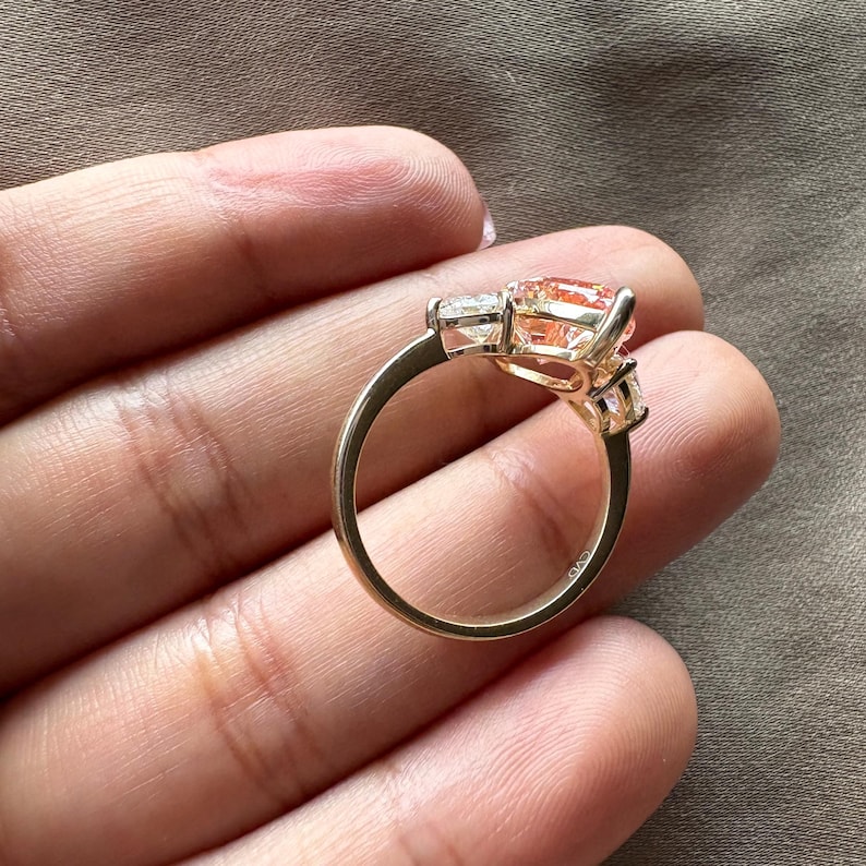 May include: A gold ring featuring a large, peach-colored gemstone flanked by two smaller, clear stones. The ring's band is a simple, polished gold, and the setting highlights the central stone's shape and color.