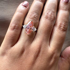 Pink marquise lab grown diamond engagement ring with heart-shaped side stones, three-stone design, 4.00 carat center diamond, elegant wedding jewelry.