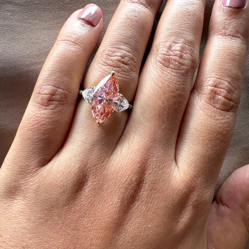 Pink marquise lab grown diamond engagement ring with heart-shaped side stones, three-stone design, 4.00 carat center diamond, elegant wedding jewelry.