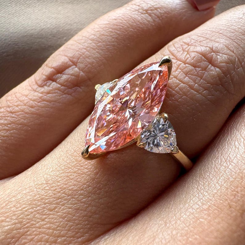 Pink marquise lab grown diamond engagement ring with heart-shaped side stones, three-stone design, 4.00 carat center diamond, elegant wedding jewelry.