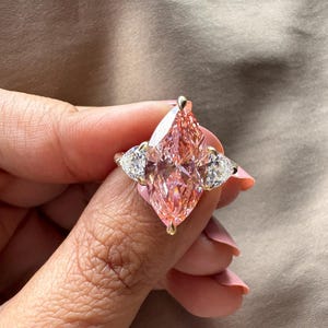 May include: A gold ring featuring a large, pink marquise-cut gemstone as the centerpiece, flanked by two smaller, pear-shaped white gemstones. The ring's setting is gold, and the gemstones sparkle.