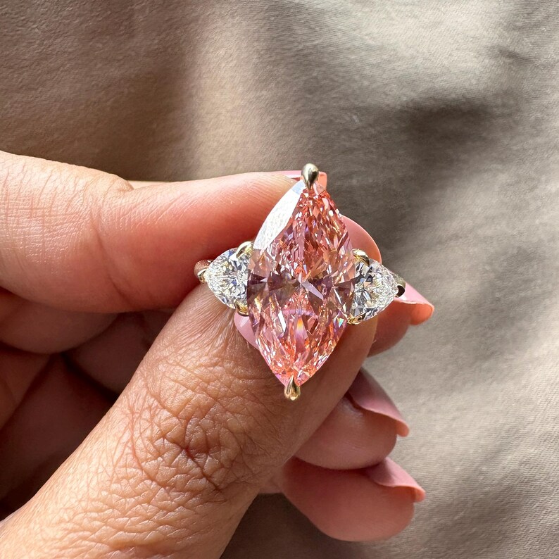 May include: A gold ring featuring a large, pink marquise-cut gemstone as the centerpiece, flanked by two smaller, pear-shaped white gemstones. The ring's setting is gold, and the gemstones sparkle.