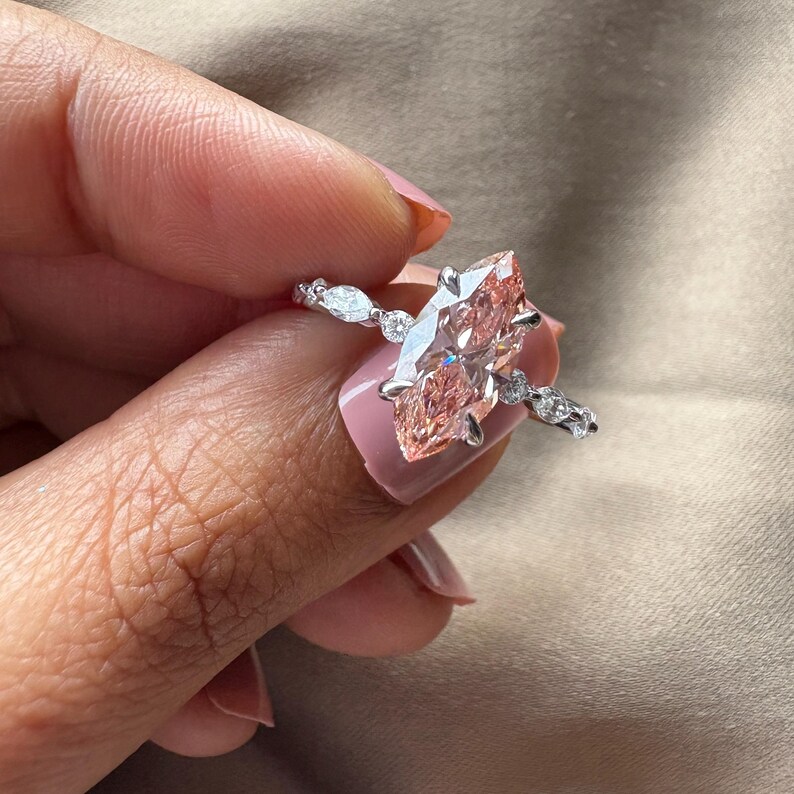 May include: A silver ring featuring a large, marquise-cut, light pink gemstone as the centerpiece. The band is adorned with smaller, clear gemstones. The ring is held up against a neutral background.