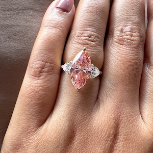 May include: A gold ring featuring a large, pink marquise-cut gemstone flanked by two smaller, white pear-shaped stones. The ring is set on a hand with a neutral-toned manicure, showcasing the jewelry's sparkle and design.