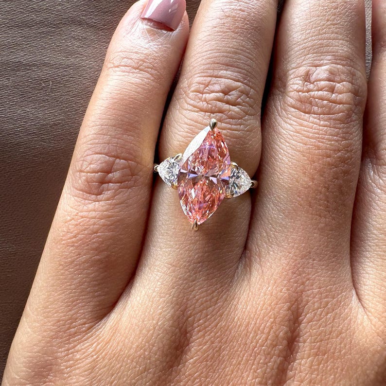 May include: A gold ring featuring a large, pink marquise-cut gemstone flanked by two smaller, white pear-shaped stones. The ring is set on a hand with a neutral-toned manicure, showcasing the jewelry's sparkle and design.