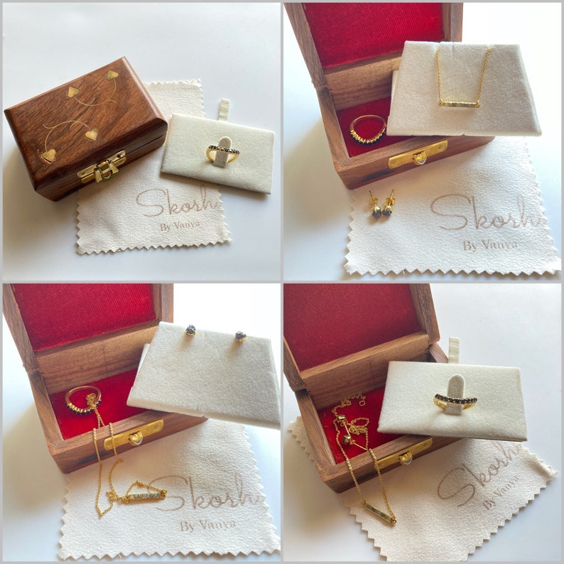 May include: A collection of gold jewelry, including rings, necklaces, and earrings, displayed in wooden boxes with red velvet lining. The jewelry is presented on white display cards and accompanied by soft, cream-colored cloths with the brand name "Skosh By Vanya".