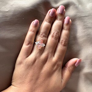 May include: A gold ring featuring a large, marquise-cut pink gemstone, flanked by two smaller, triangular white gemstones. The ring is set on a hand with pink nail polish, against a neutral background.