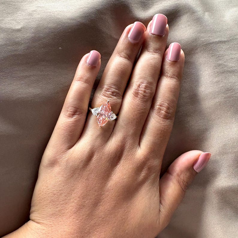 May include: A gold ring featuring a large, marquise-cut pink gemstone, flanked by two smaller, triangular white gemstones. The ring is set on a hand with pink nail polish, against a neutral background.