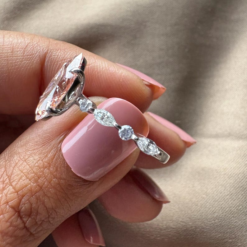 May include: A silver ring with a large, light peach-colored, marquise-cut gemstone as the centerpiece. The band features smaller, clear gemstones in a delicate design. The ring is held up against a background of a person's skin and a neutral-toned surface.