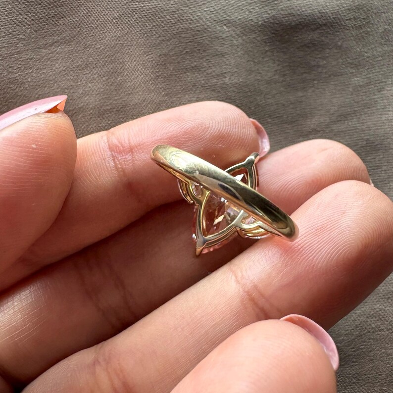 May include: A gold ring with a large, light pink, pear-shaped gemstone. The ring's band is a simple, polished gold, and the gemstone is set in a delicate, openwork setting. The ring is shown from a side angle.