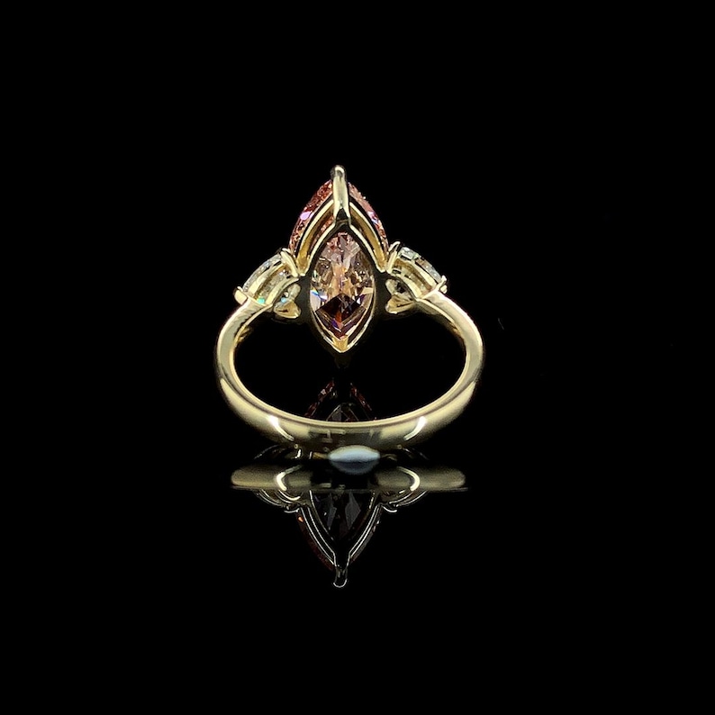 May include: A gold ring with a marquise-cut, peach-colored gemstone as the centerpiece. The ring features smaller, clear gemstones on either side of the main stone, set against a black background.