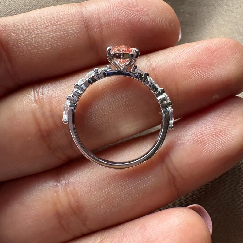 May include: A silver ring with a peach-colored gemstone center and smaller clear stones along the band. The ring is held in a hand, showcasing its intricate design and sparkle. The ring is a piece of jewelry.