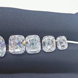 May include: Five clear, colorless, cushion-cut gemstones arranged in a row from largest to smallest. Each stone features a faceted surface that reflects light, creating a brilliant, sparkling appearance against a dark, smooth fabric background.