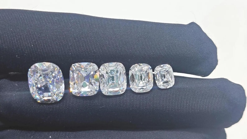 May include: Five clear, colorless, cushion-cut gemstones arranged in a row from largest to smallest. Each stone features a faceted surface that reflects light, creating a brilliant, sparkling appearance against a dark, smooth fabric background.