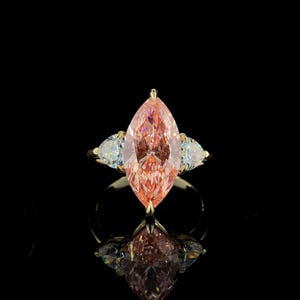 May include: A gold ring featuring a large, marquise-cut pink gemstone flanked by two smaller, pear-shaped white gemstones. The ring is set against a black background, with its reflection visible below.