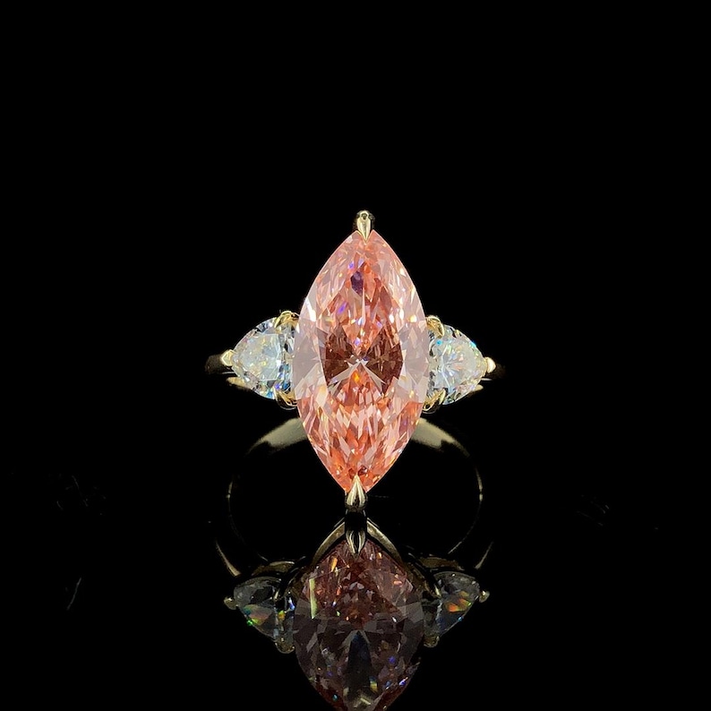 May include: A gold ring featuring a large, marquise-cut pink gemstone flanked by two smaller, pear-shaped white gemstones. The ring is set against a black background, with its reflection visible below.