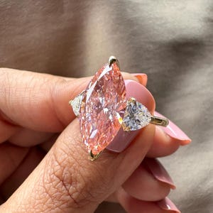 May include: A gold ring featuring a large, marquise-cut pink diamond as the centerpiece, flanked by two smaller, triangular white diamonds. The ring's setting is gold, and the diamonds sparkle.