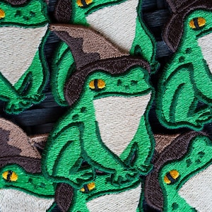 May include: Close-up of several green frog embroidered patches with brown felt hats. The frogs have white felt bellies and yellow eyes.