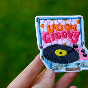 May include: A blue and pink embroidered patch of a vintage record player with the text "Let's Get Groovy!"