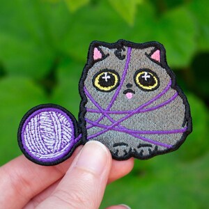 May include: An embroidered grey cat patch with large eyes and a pink tongue, tangled in purple yarn. The cat is holding a ball of purple yarn.