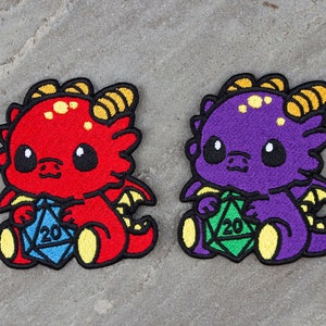 Peut inclure: Deux patchs brodés de mignons dragons de dessin animé tenant des dés. Le dragon rouge tient un dé bleu à 20 faces, et le dragon violet tient un dé vert à 20 faces.