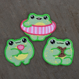 May include: Three green frog embroidered patches. One frog is holding a pink flower, another is holding a brown cookie, and the third is wearing a pink and white striped life preserver.