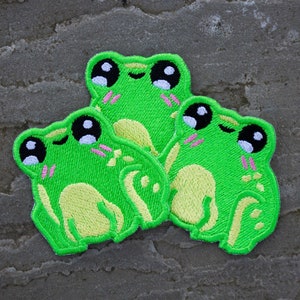 May include: Three green embroidered frog patches with black eyes and pink cheeks. The frogs are smiling and have a yellow belly.
