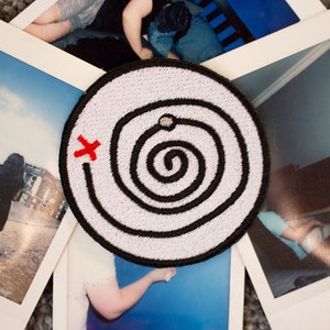 Può includere: Toppa ricamata bianca con un motivo a spirale nero e una X rossa sul lato sinistro. La spirale ha un piccolo punto grigio al centro.