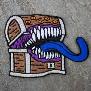 Può includere: Una toppa ricamata che rappresenta uno scrigno del tesoro con la faccia di un mostro e una lingua blu che sporge. Il mostro ha denti bianchi e occhi arancioni.