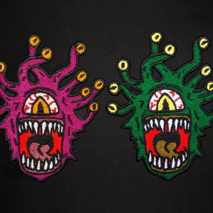 May include: Two embroidered patches depicting a mythical creature with multiple eyes and a large mouth. The creature on the left is pink with yellow eyes, while the creature on the right is green with yellow eyes.