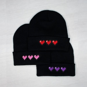 Può includere: Tre berretti di lana neri con disegni di cuori pixelati. Il berretto superiore ha tre cuori rossi, il berretto intermedio ha tre cuori rosa e il berretto inferiore ha tre cuori viola.