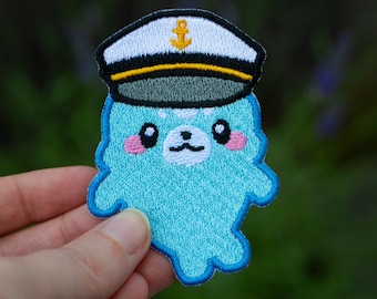 May include: A blue embroidered patch of a cartoon ghost wearing a white and black sailor hat with an anchor on the front. The ghost has a cute face with black eyes, pink cheeks, and a white nose.
