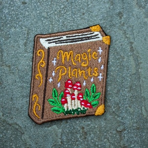 May include: Embroidered patch of a book with the title "Magic Plants" and illustrations of mushrooms and leaves. The book is brown with gold accents.