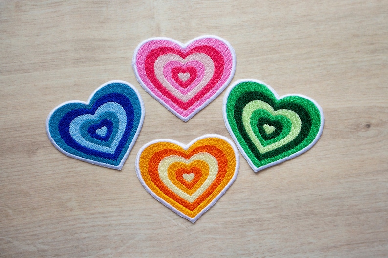 May include: Four embroidered heart-shaped patches in different colours. The hearts are layered with concentric circles of colour, creating a rainbow effect. The colours are pink, blue, green, and orange.
