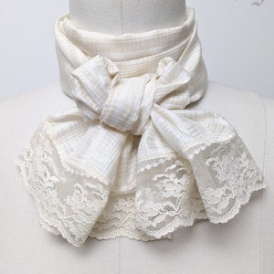May include: A white cravat with a checkered pattern and a lace trim. The cravat is tied in a bow and is displayed on a white mannequin. The text "Rare Dandy" is visible in the upper right corner of the image.