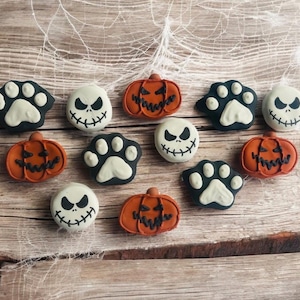 May include: A collection of Halloween-themed cookies arranged on a wooden surface. The cookies feature designs of jack-o'-lanterns, paw prints, and spooky faces in black, white, and orange. The background includes a spiderweb.