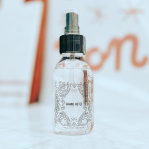 May include: A clear glass spray bottle with a black cap and pump. The label features the words "GRAND HOTEL" in a decorative frame. The bottom of the label reads "DOLE NICKS CANDLE CO."