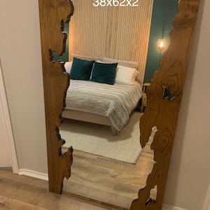 May include: A large, rectangular mirror with a unique, natural wood frame. The frame has a rustic, live edge design with a dark brown stain. The mirror reflects a bedroom with a bed, pillows, and a nightstand.