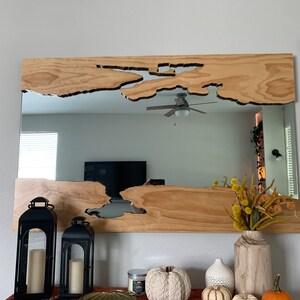 May include: A wooden mirror with a unique, irregular shape cut out of the wood. The mirror is hanging on a wall above a mantle with candlesticks, a vase of dried flowers, and pumpkins.