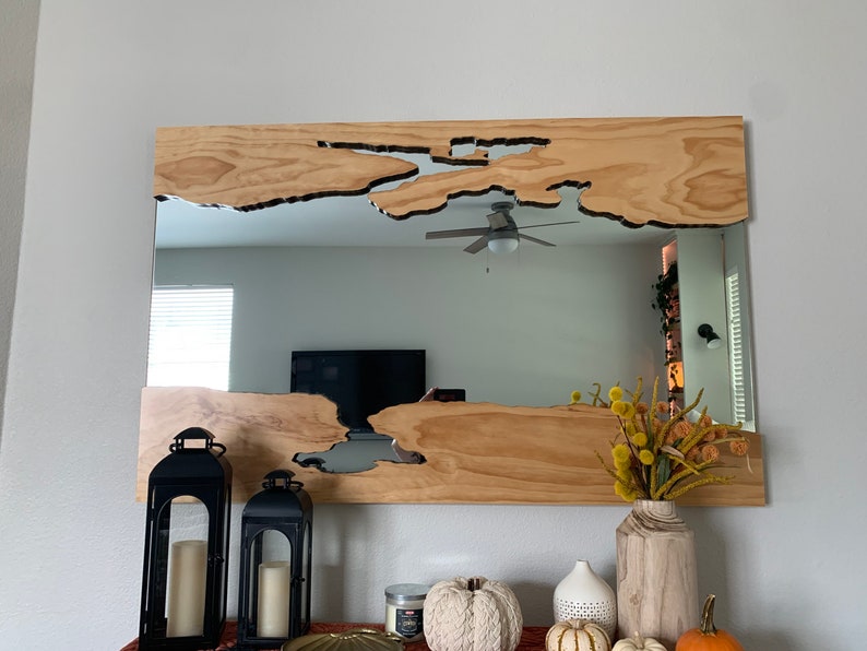 May include: A wooden mirror with a unique, irregular shape cut out of the wood. The mirror is hanging on a wall above a mantle with candlesticks, a vase of dried flowers, and pumpkins.