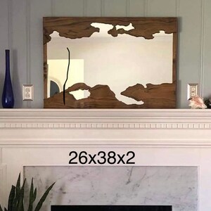 May include: A wooden framed mirror with a unique, irregular shape cut out of the wood. The mirror is hanging above a white fireplace mantel with a marble surround. The text "26x38x2" is printed below the mirror.