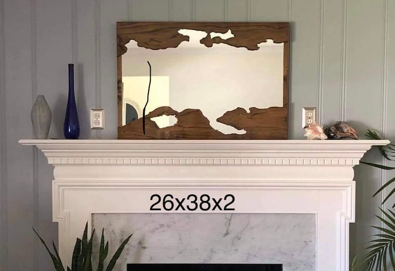 May include: A wooden framed mirror with a unique, irregular shape cut out of the wood. The mirror is hanging above a white fireplace mantel with a marble surround. The text "26x38x2" is printed below the mirror.