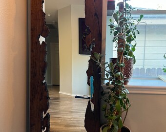 May include: A large, dark brown wooden mirror with an irregular, natural edge frame. The mirror is reflecting a room with hardwood floors and a painting on the wall. A potted plant with green leaves is in the foreground.